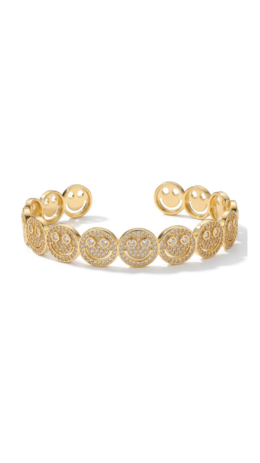 A gold-plated open-cuff bangle featuring a repeating pattern of pavé crystal smiley faces on a white background.
