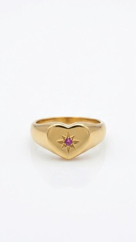 A polished 18K gold-plated heart signet ring with a starburst engraving and a small pink crystal at the center on a white background.