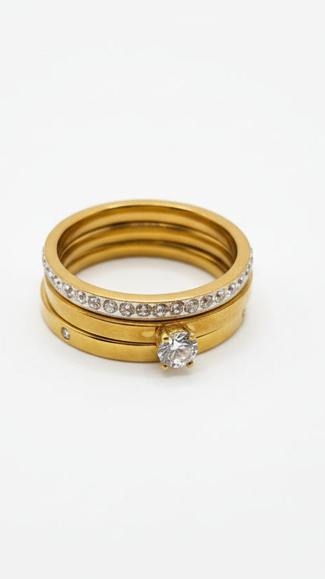 A stack of three 18K gold-plated rings: one with a large round solitaire crystal, one with micro-pave crystals, and one plain polished band.