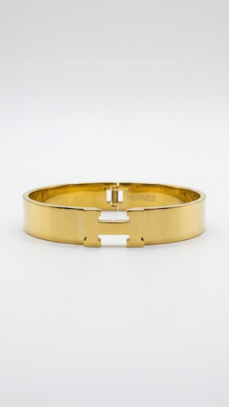 A wide, polished 18K gold-plated hinged bangle with a central H motif on a model's wrist.